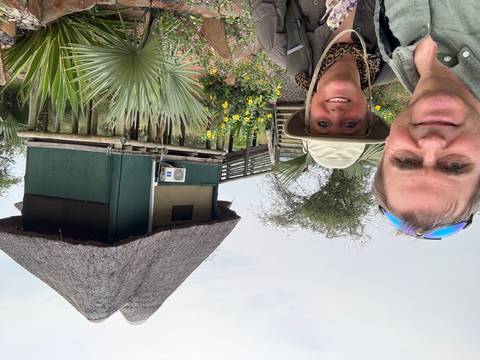 Two people posing in front of a stilted structure with a thatched roof.