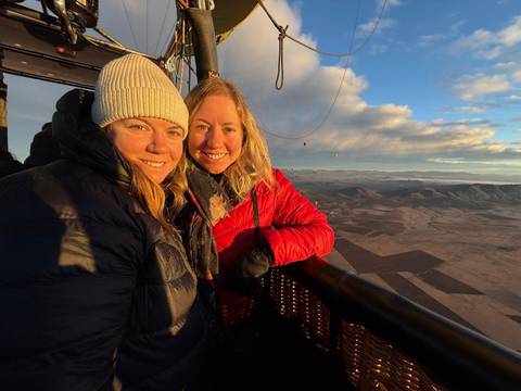 Two people pose in a hot air balloon basket with a sunrise view.