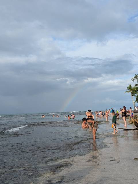 Crowded beach with people enjoying the water and a rainbow in the sky.