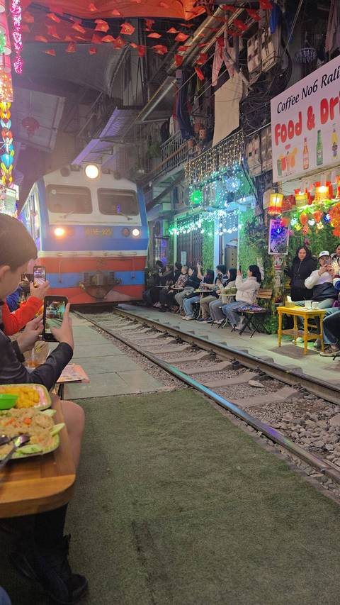       Train moving through a narrow street with people taking photos.
  