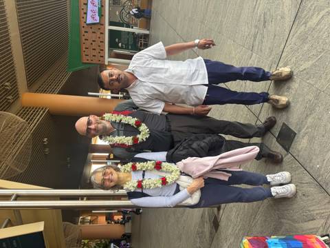       Three people standing, two wearing garlands in a public space.
  