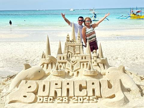       Two people pose with a large sand sculpture on a beach.
  