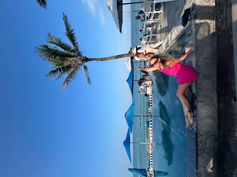 Woman in pink dress posing by a pool with the ocean and palm trees in the background.