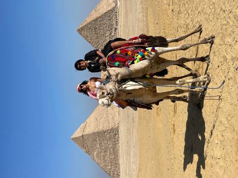 Two people on camels with pyramids in the background.