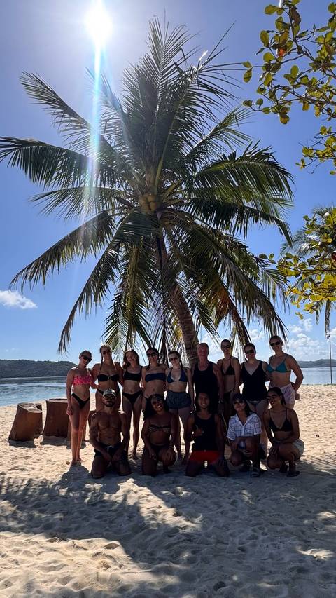 Group of people standing together under a palm tree on a sunny beach.