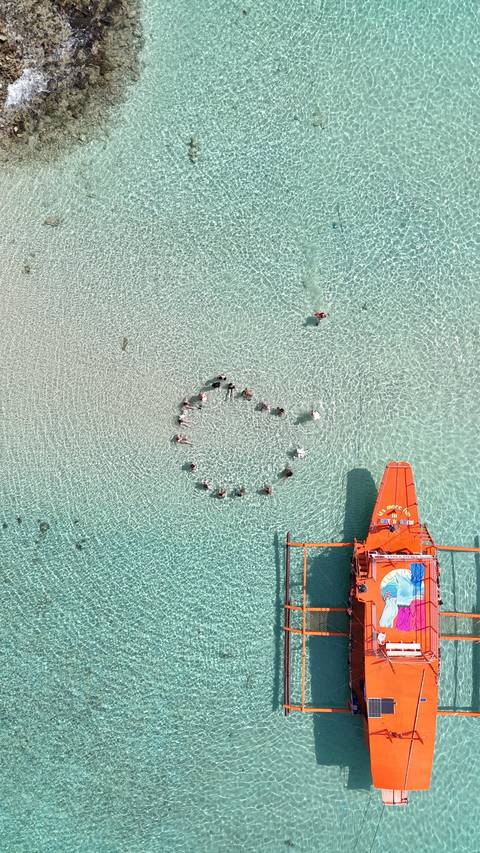 Overhead view of people forming a circle in a turquoise shallow sea with a boat nearby.