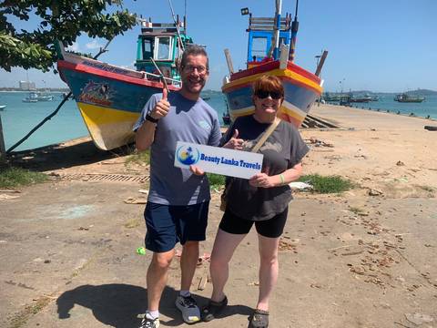      Two people posing with a 'Beauty Lanka Travels' sign by colorful boats at a dock.
  