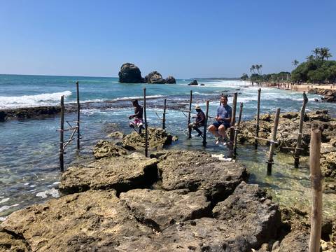       Three people sitting on stilts above the rocky coastline, with clear blue water and distant rocks.
  