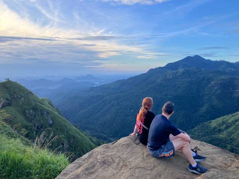       Two people sitting on a rock, overlooking a mountainous landscape with a colorful sky.
  