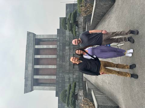 Three people posing in front of a large stone building with columns and a red wall.