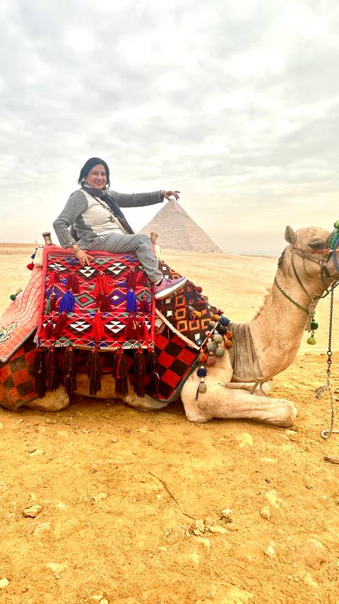 Person on a decorated camel with a pyramid in the sandy background.