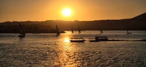       Silhouettes of boats sailing on a river at sunset with golden reflections.
  