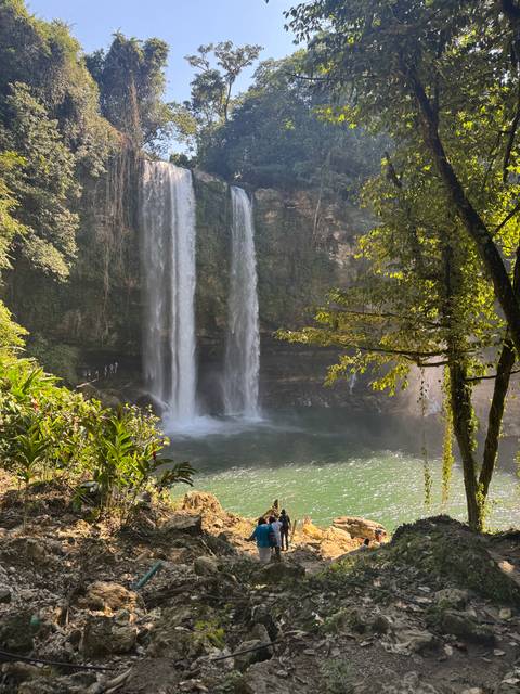 Two tall waterfalls with people near the base, surrounded by dense forest.