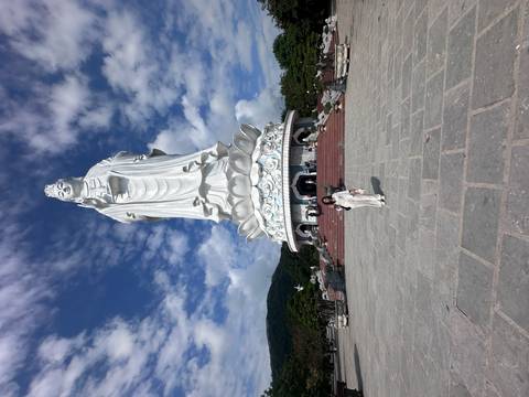       A large statue of a deity with a person standing in front under a blue sky.
  