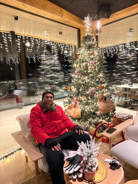 Person in winter clothing sitting by a decorated Christmas tree.