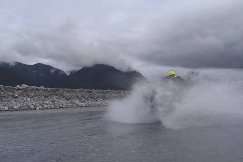       Person on a quad bike riding through a wet terrain with mountains in the background.
  