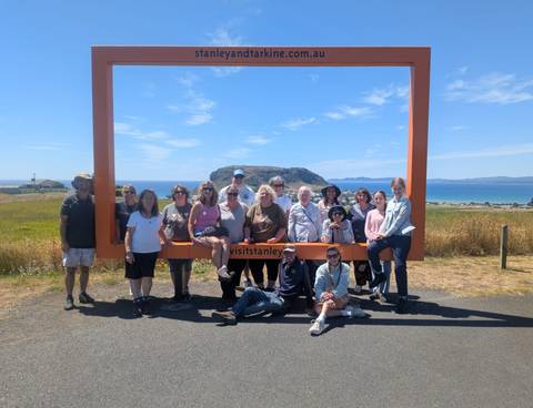       Large group posing by a large yellow frame with a scenic ocean view.
  