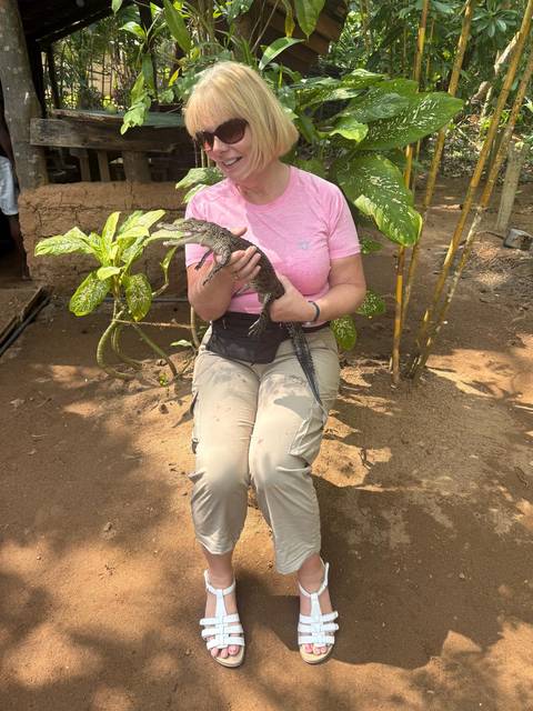       A person holding a small crocodile or reptile, seated outdoors with plants in the background.
  