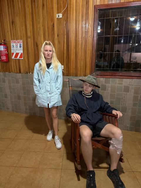       Two people indoors, one standing, one sitting on a chair, wooden wall behind.
  