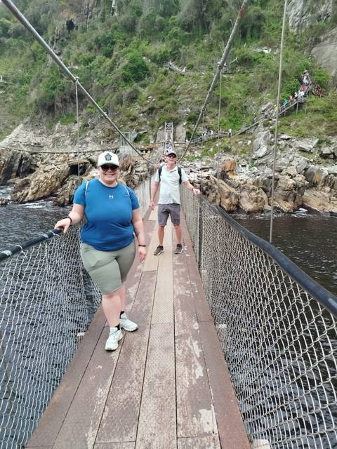       Two people crossing a suspension bridge over a river in a lush area.
  