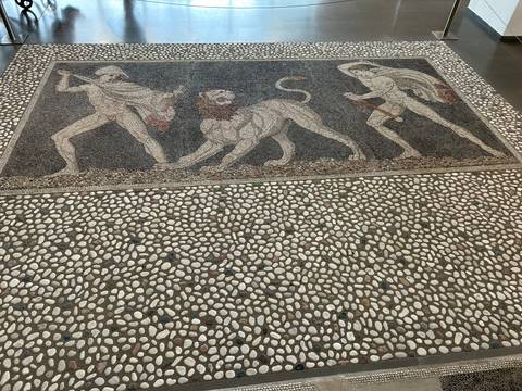 Floor mosaic depicting figures and a lion in a museum.