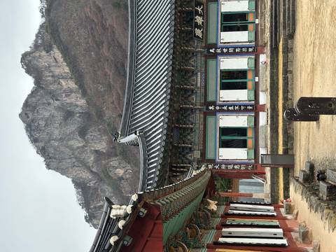       Historic Korean temple hall decorated with colorful patterns set below a rugged forested mountain cliff.
  
