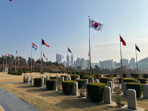       Flag-lined cemetery with gravestones and distant skyline of Busan rising through light haze.
  