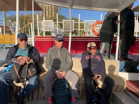 Three older men sitting on a boat deck under sunlight.
