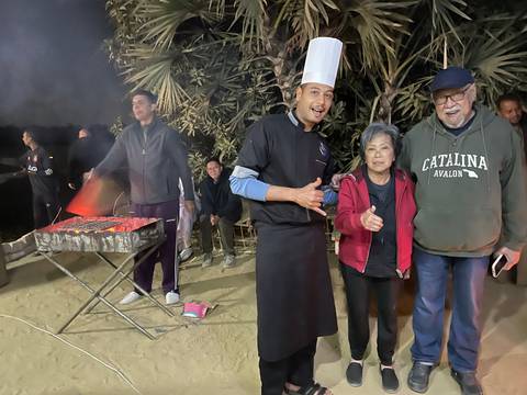       Casual night-time barbecue scene with a chef tending coals and travellers posing for a photo.
  