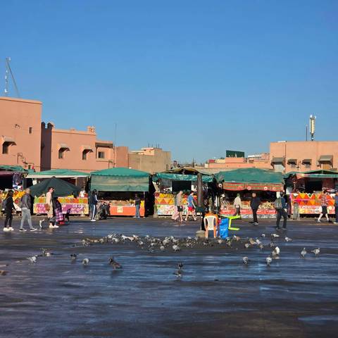       Busy market with people walking and colorful stalls under clear blue sky.
  