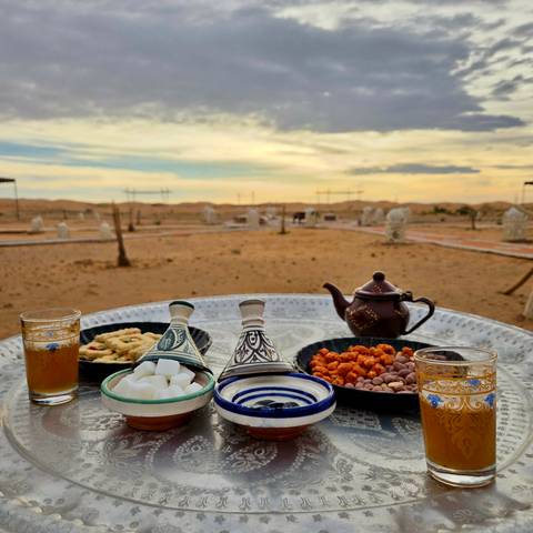       Table with traditional Moroccan tea setup in a desert setting at sunset with tents in the background.
  