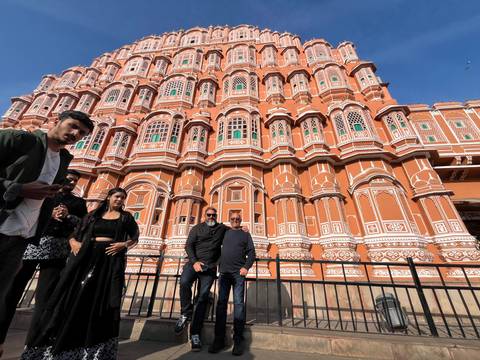       Visitors pose below the ornate pink lattice façade of Jaipur's Hawa Mahal under a blue sky.
  