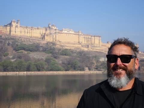       Smiling traveler with sunglasses stands before the sprawling yellow walls of Amber Fort across the lake.
  