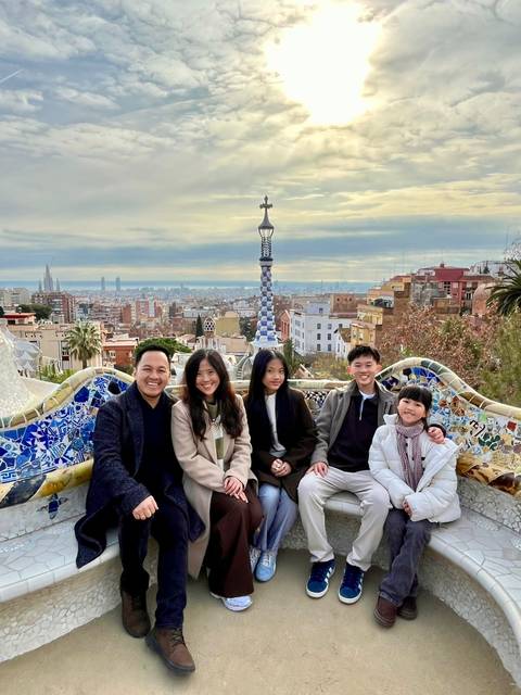       Family posed on colourful mosaic bench overlooking Barcelona skyline in Park Güell.
  