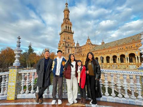       Group photo on ornate bridge railings with the grand Plaza de España’s towered building in Seville behind.
  