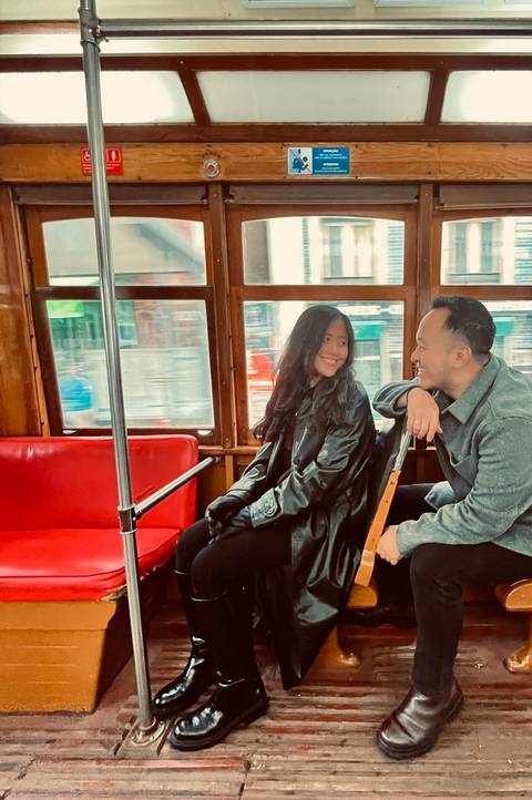       Couple smiling at each other while sitting inside a vintage tram with wooden interior and city view outside.
  