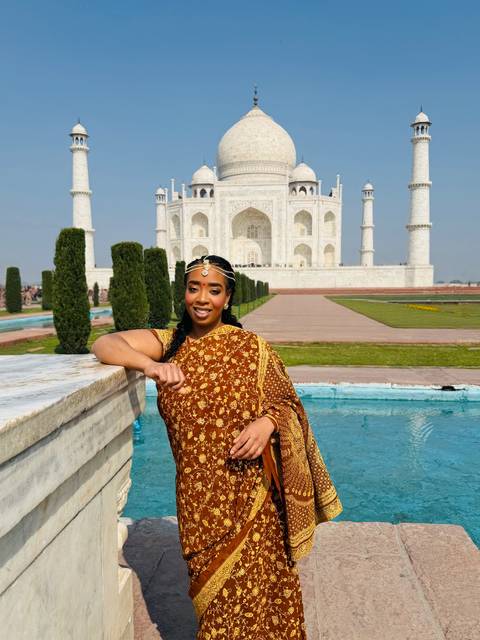       Woman leaning on marble railing with Taj Mahal’s white domes and minarets reflected in blue pool.
  