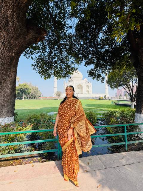      Woman in sari framed by leafy trees with distant view of the Taj Mahal across lush grounds.
  