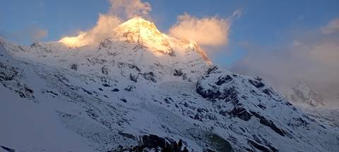       First light hits a snow-clad peak in the Annapurna range, glowing above clouds.
  