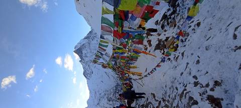       Prayer flags flutter against a snowy mountain backdrop with trekkers gathering nearby.
  