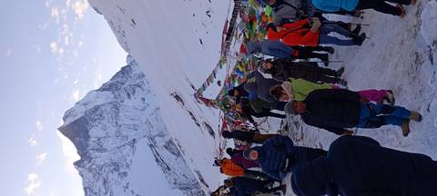       Large group of trekkers at snowy Annapurna Base Camp surrounded by colourful prayer flags and peaks.
  