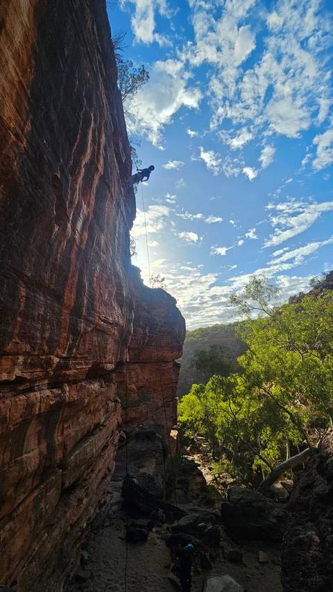       Sun-lit sandstone cliff towering over a lush green valley with blue sky and scattered clouds; a rope hangs down the rock face.
  