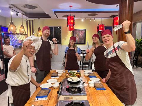       Cooking class participants wearing aprons and chef hats pose with knives around a table set with Vietnamese ingredients.
  