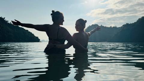      Two women standing waist-deep in calm bay waters with forested karst cliffs behind, arms outstretched toward the sun
  
