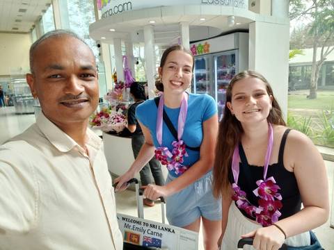       Smiling guide and two female travelers with orchid garlands take a selfie in an airport arrivals hall.
  