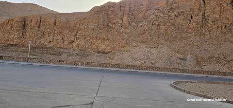       Empty asphalt lot bordered by a rocky desert cliff under a pale sky.
  