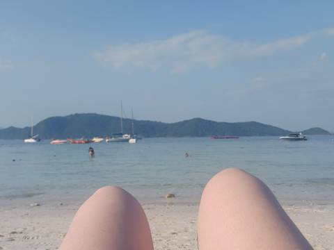       Beach view from the perspective of someone lying down with knees in foreground, boats and islands beyond.
  