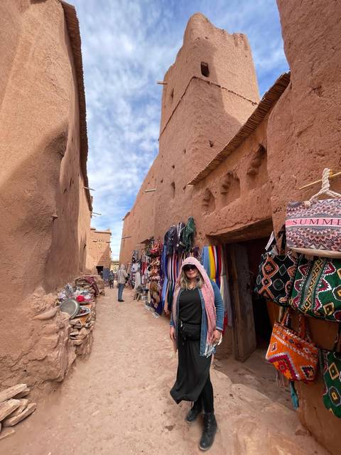       Narrow adobe alley lined with colorful textiles and market stalls in a Moroccan ksar.
  