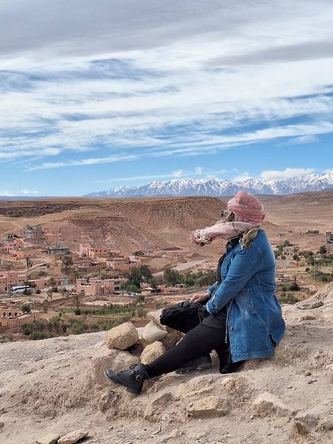       Traveller in a headscarf gazes over a desert village toward snow-capped Atlas Mountains.
  