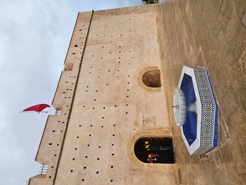       Historic Moroccan fort wall with tiled fountain and national flag fluttering above the entrance.
  